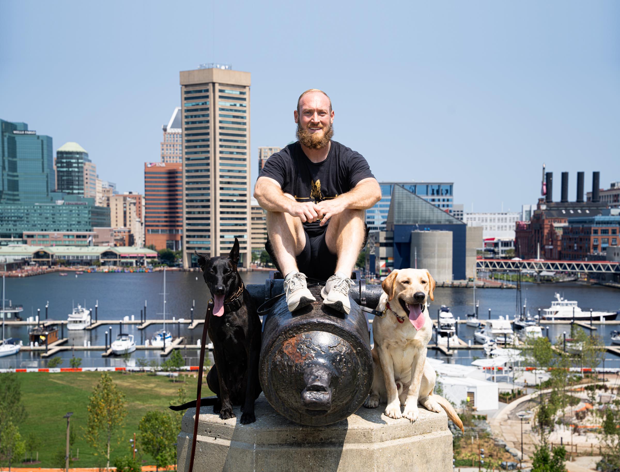 Nick Gallina sitting on a cannon above Baltimore&rsquo;s Inner Harbor, a black shepherd and a yellow Labrador holding a sit either side of him